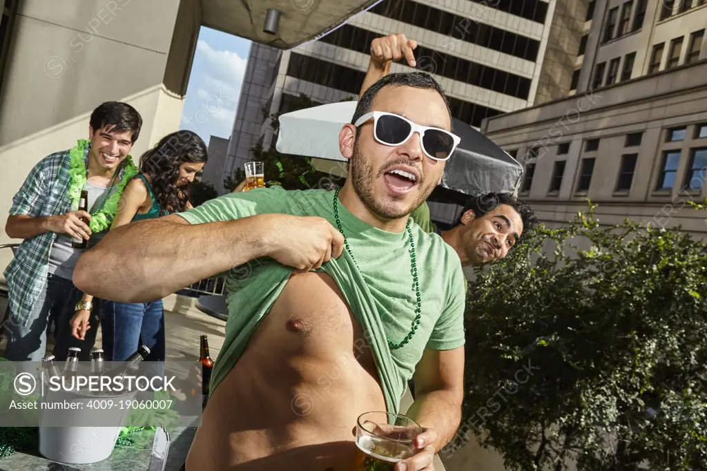 Man pulling up shirt and friends laughing while drinking and partying during St Patrick's Day. 
