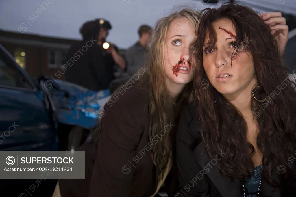 Injured young women sitting in street  with scene of an automobile accident behind them