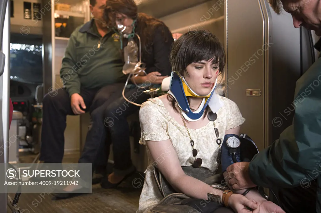 Paramedic attending to injured woman wearing a neck brace sitting on back of open ambulance, while her friend is getting oxygen in the back with another EMT 