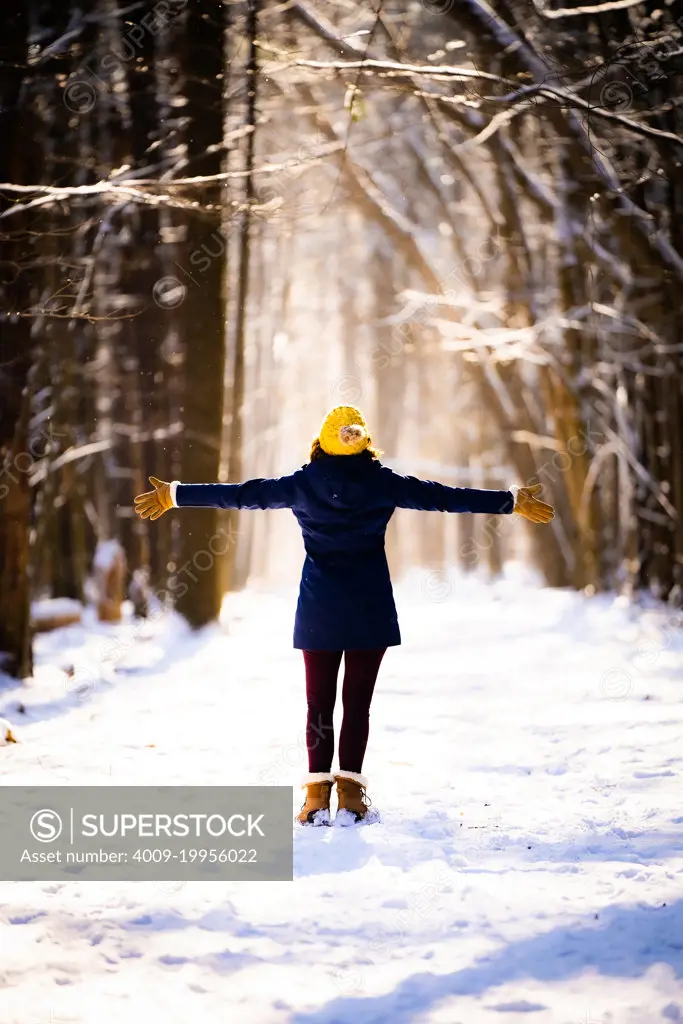 A woman spreading her arms out and looking up during a hike through the woods in the Winter at sunset.