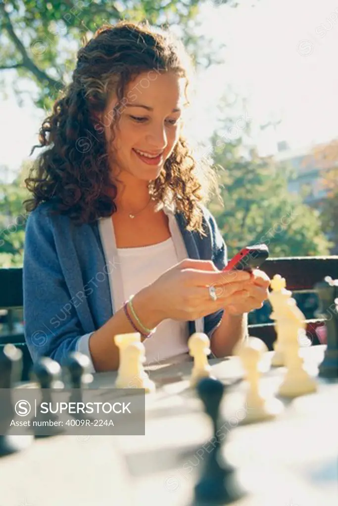Young woman text messaging on mobile phone while playing chess