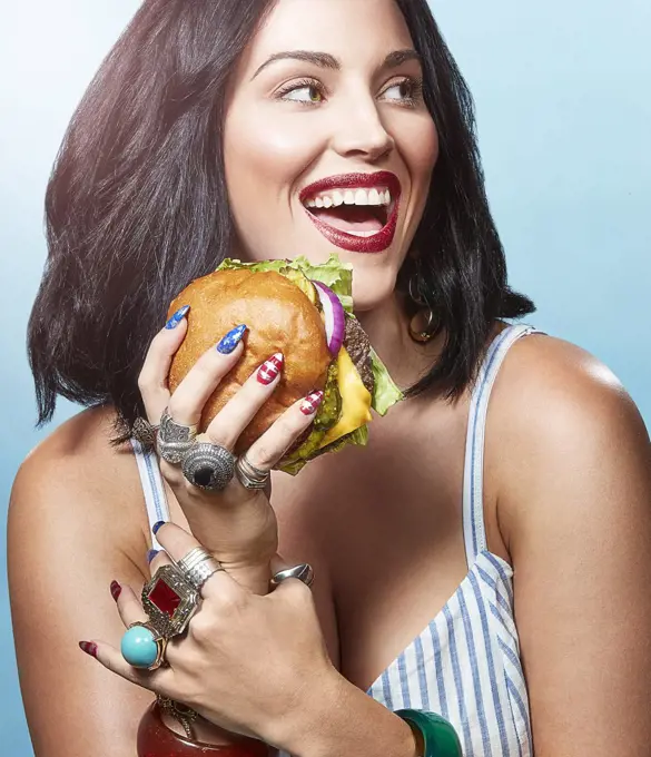 A woman in front of a blue background eating a burger