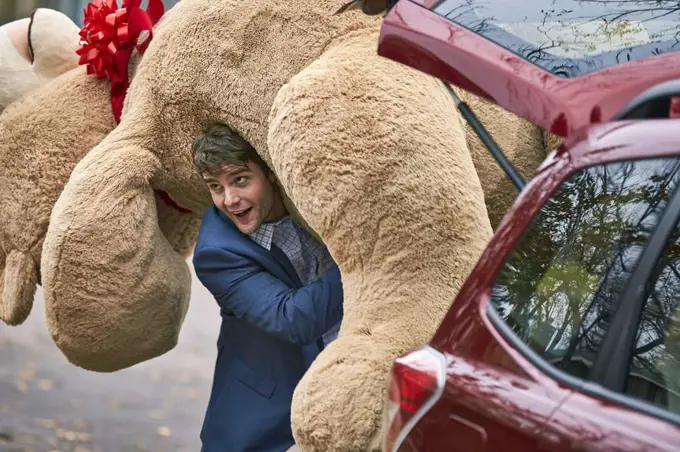 3/4 MCU of a young man grabbing a large teddy bear from the trunk of his car, throwing it over his shoulder and walking off camera.
