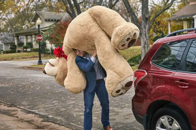 Wide shot of a man carrying a large teddy bear over his shoulder.