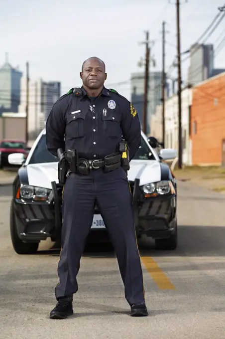 Portrait of Police officer Standing in street in front of squad car  looking towards camera with arms behind his back 