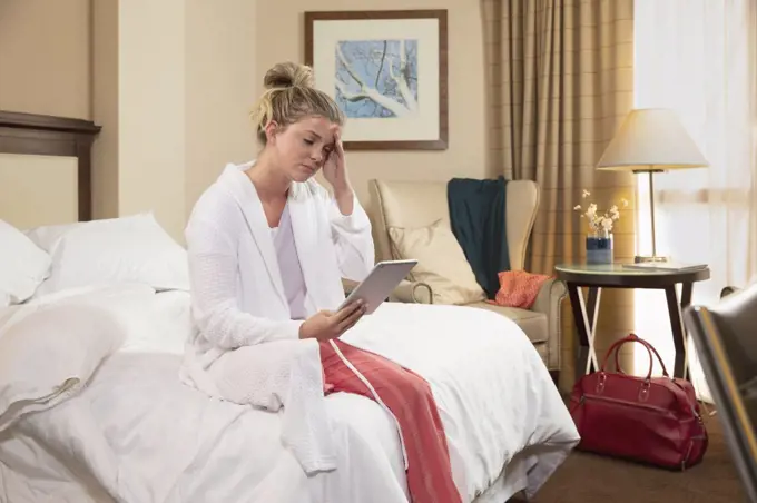 Young Caucasian woman with headache wearing bathrobe in a hotel room, sitting on edge of bed using tablet device to connect with doctor