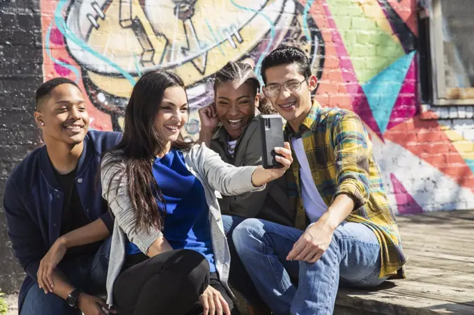 Group of young co-workers hanging out in front of graffitied wall taking selfie with mobile phone 