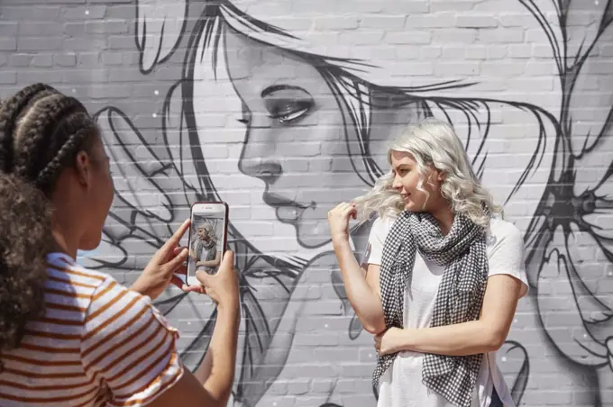 Two young women in front of graffitied wall using mobile phone to take portrait 