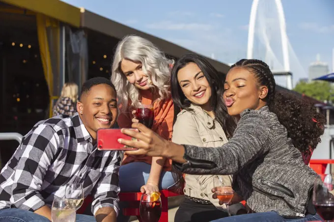 Group of young co-workers hanging out on outdoor patio having drinks for happy hour, woman taking group photo with mobile phone 