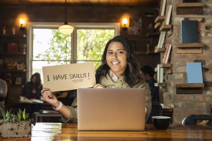 Portrait of young woman sitting at table in coffee shop bookstore with laptop computer , holding hand made sign that reads ÒI have skills