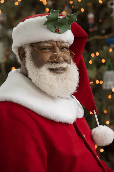 An African American man dressed as Santa Claus sitting in front of a Christmas tree looking camera smiling. 