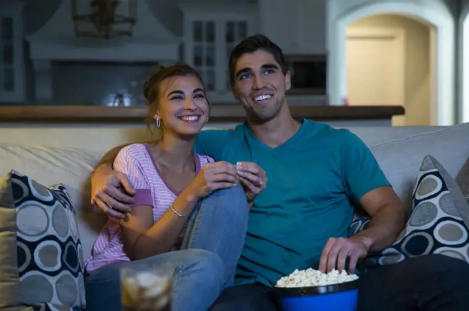 Young couple sitting on couch watching a movie at home with a bowl of popcorn 