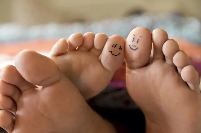 Close-up of bare feet sticking out from covers while couple lays in bed, with smiley faces drawn on big toes