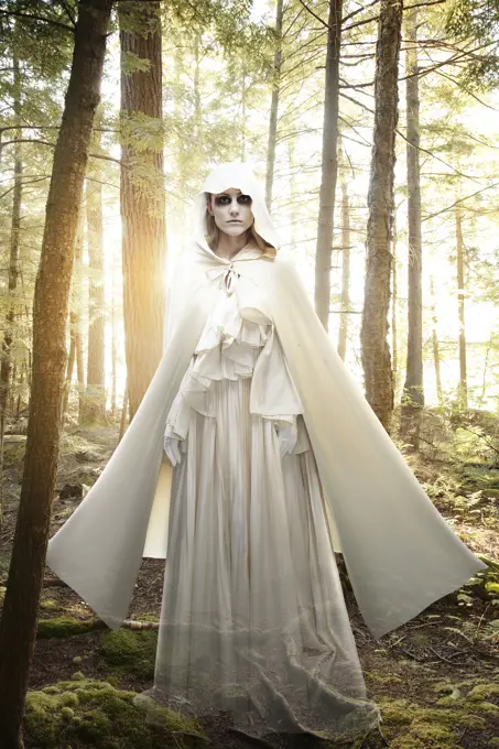 Female ghost with black circles around her eyes, dressed all in white standing in a forest at sunrise
