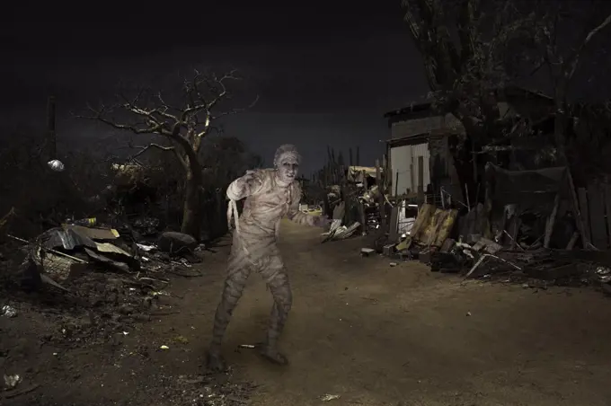 Mummy walking on a dirt path near an abandoned building surrounded by trash at night
