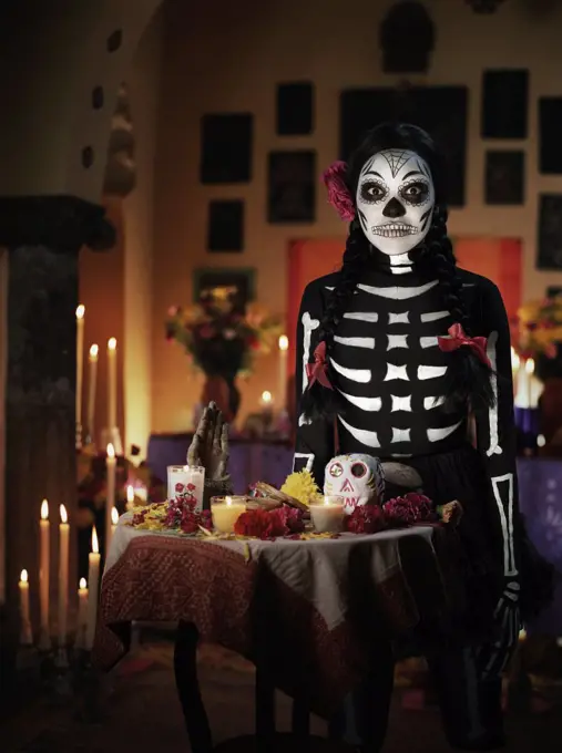 Female skeleton with ribbons and a flower in her hair standing behind a Dios de los Muertos altar