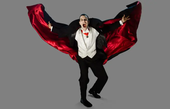 Themed portrait of a vampire on a gray background lunging towards camera with cape flying out behind him, fangs out and mouth open