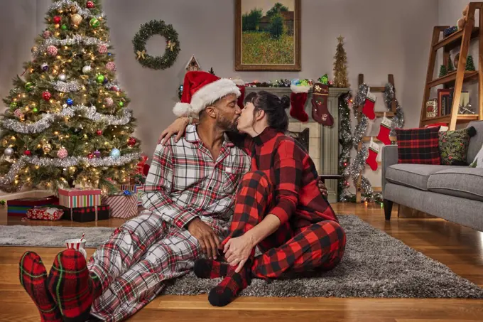 Romantic young diverse couple kissing while sitting in living room by Christmas tree.