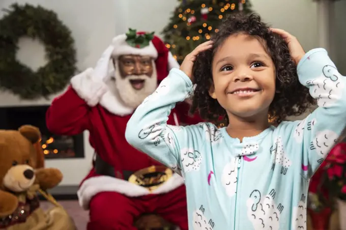 Young child in living room with a Christmas background and Santa mimicking her moves behind her. Black Santa.