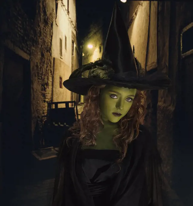 Young girl dressed as witch standing in front dark alley 