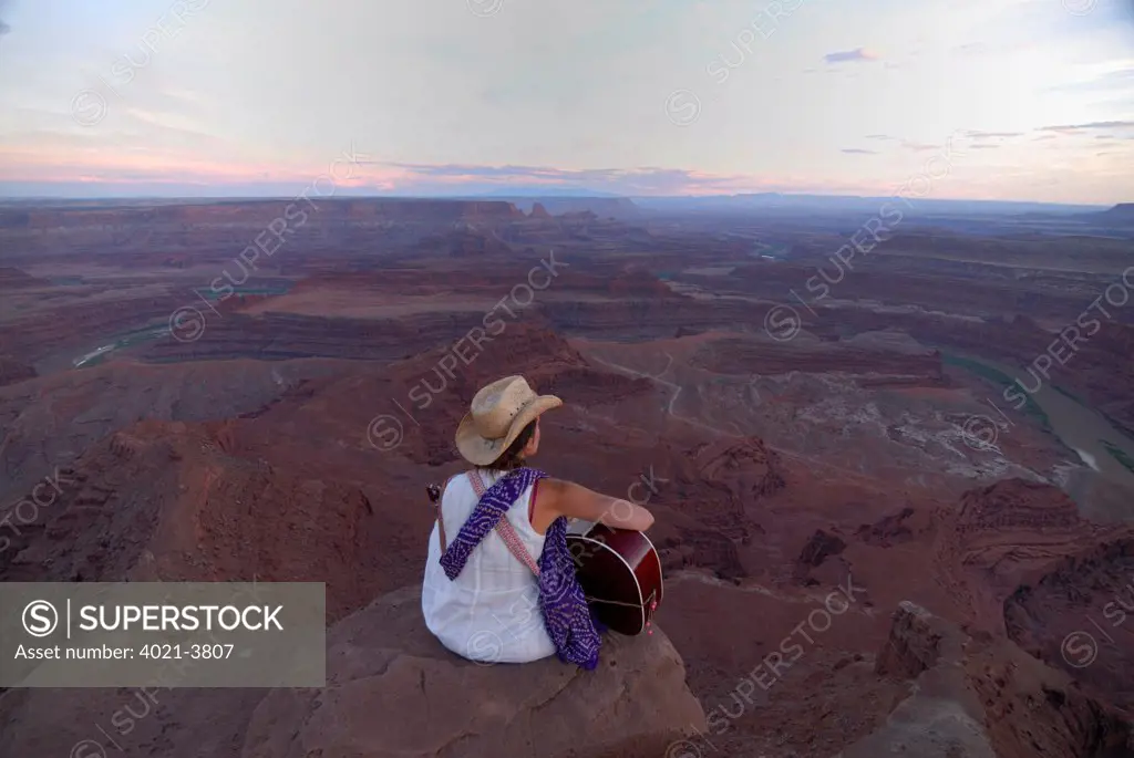 Woman with cowboy hat playing the guitar and singing a song while sitting on the edge of a canyon at Dead Horse Point State Park near Moab, Utah, USA