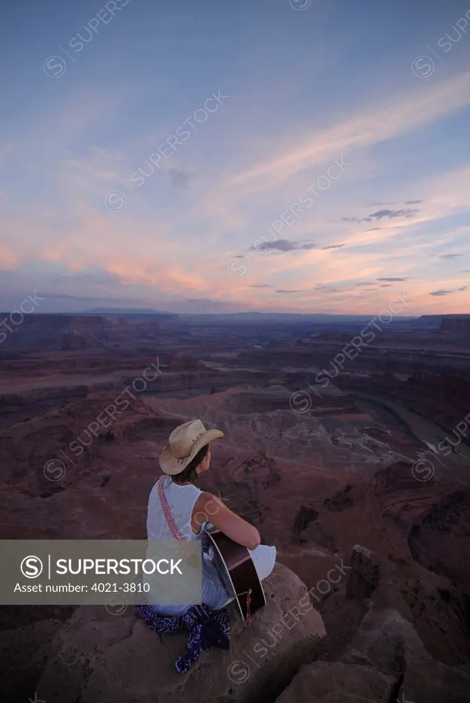 Woman with cowboy hat playing the guitar and singing a song while sitting on the edge of a canyon at Dead Horse Point State Park near Moab, Utah, USA