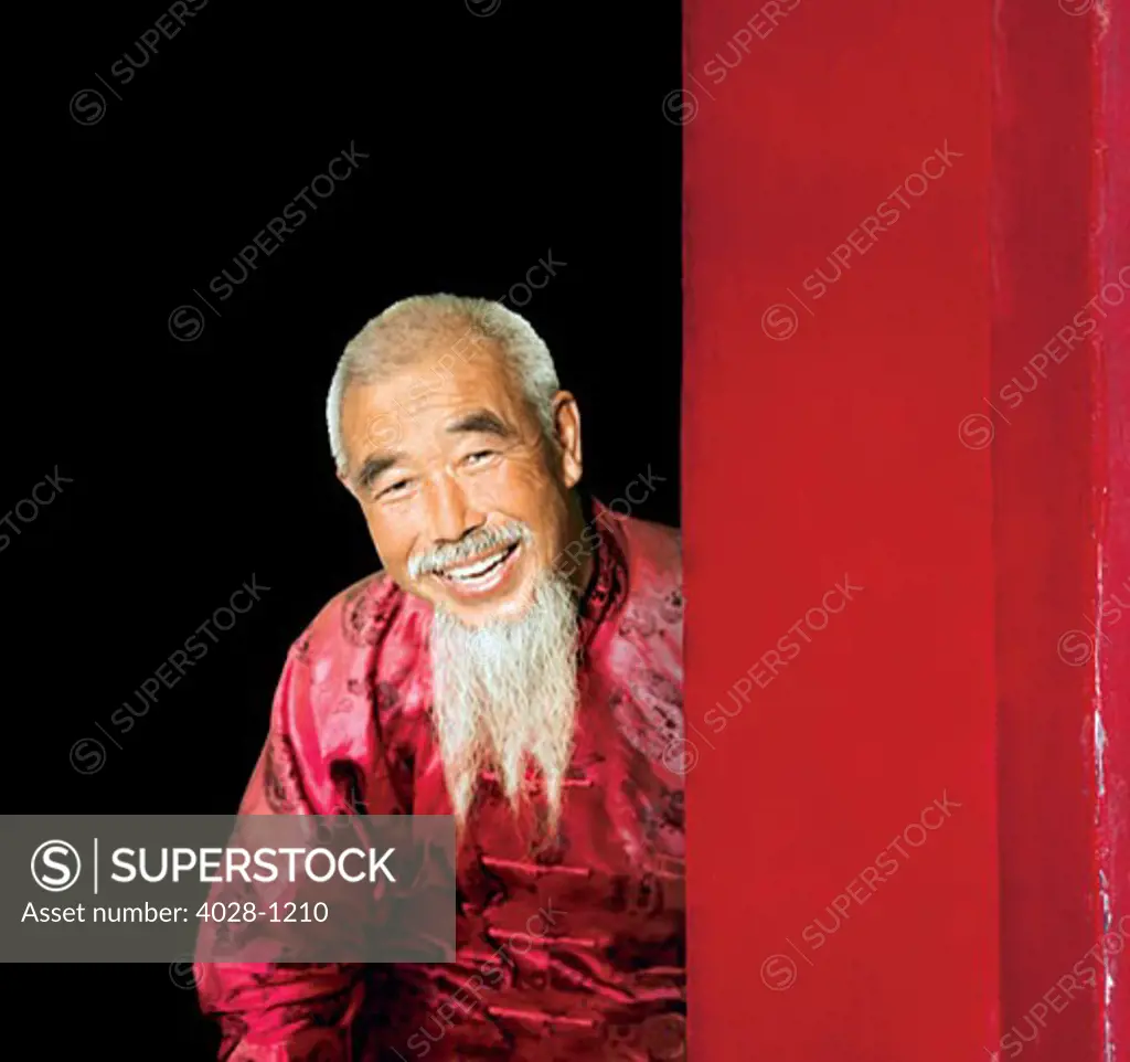 China, Beijing, An old Chinese man dressed in traditional clothes with a long white beard peers around a doorway