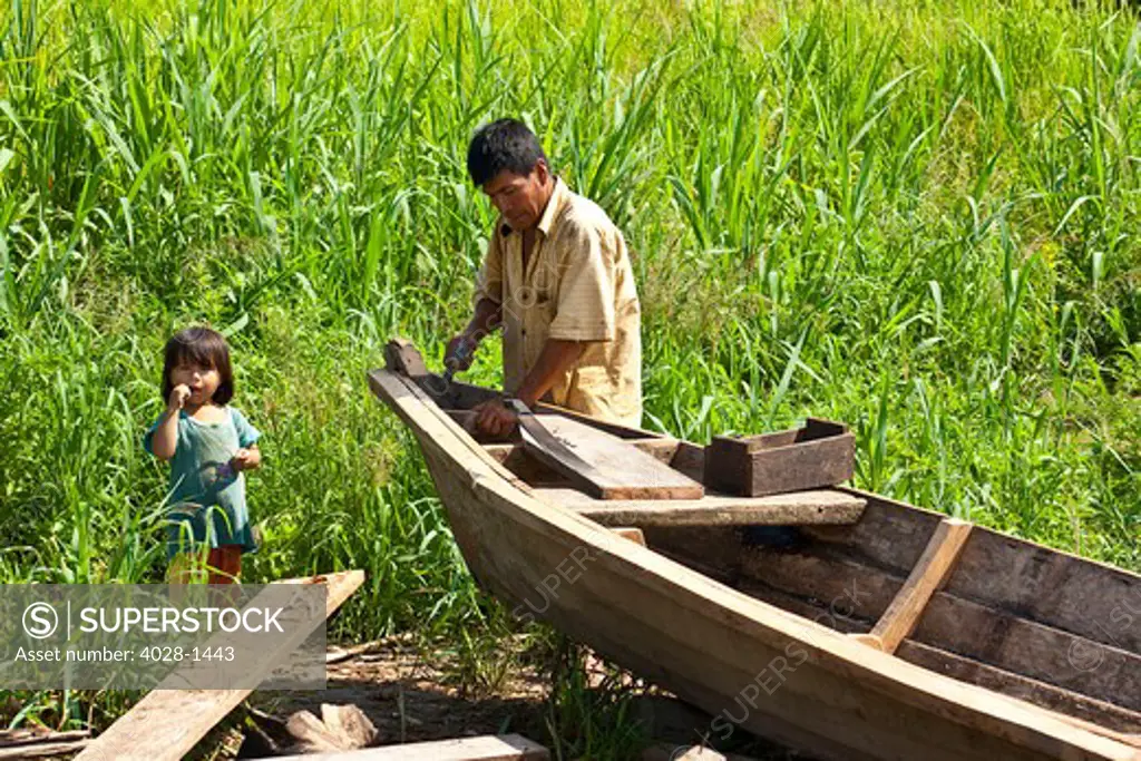 Iquitos, Peru, Amazon Jungle, A Yagua Tribe man builds a canoe along the Amazon River as his little girl looks on.