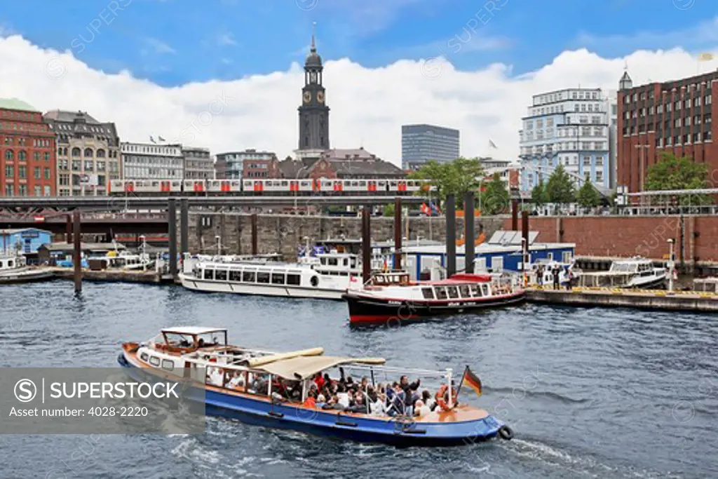 Hamburg, Germany, A tour boat moves along the waterfront warehouses and lofts in the Speicherstadt warehouse district with the Tower of St Michaelis church and a  subway train