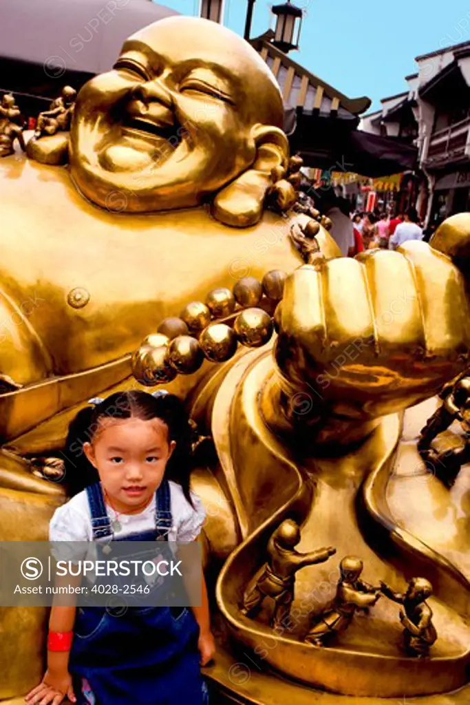 China, Hangzhou, Old Town, A young Chinese girl, child, sits at the base of a brass Buddha dedicated to children.