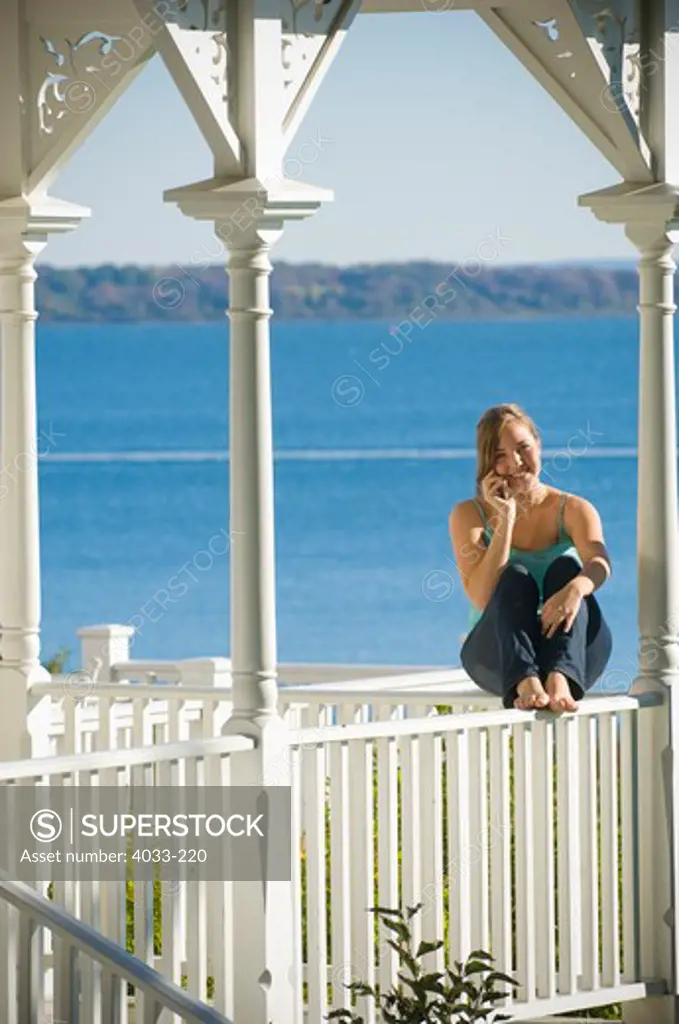 Young woman sitting on a porch railing and talking on a mobile phone, Jamestown, Rhode Island, USA