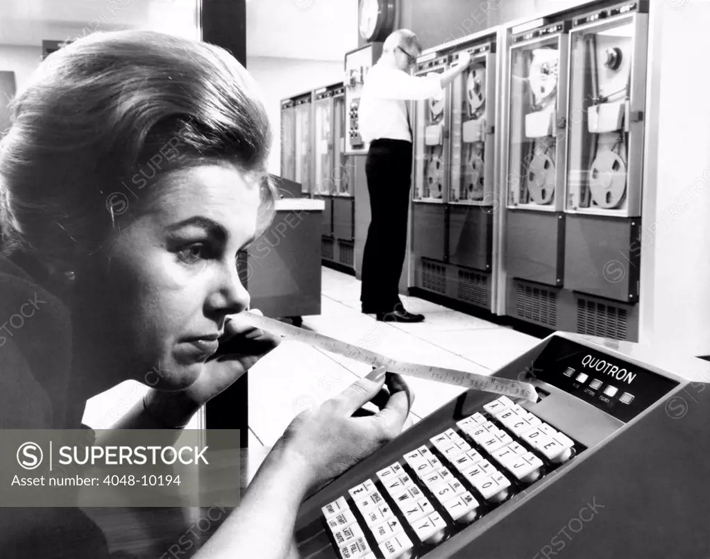 Woman holds tape with the latest stock prices printed by the Quotron II. It can recall on command the last quote of a given stock from the centers computing equipment in the background. This unit is at Scantlin Electronics Inc. 60 Broad Street office in NYC. Oct. 30, 1964.
