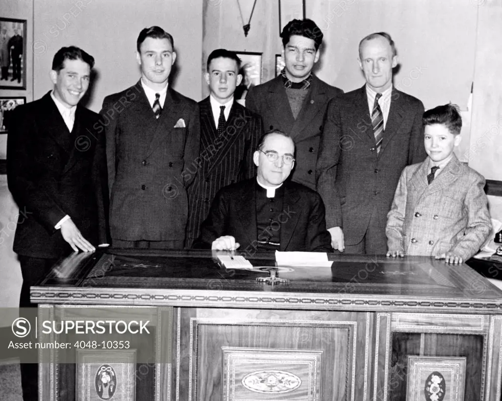 Father Edward Flanagan proudly sits on his desk made by Boys Town citizens over a period of three years. The boys who worked on the desk and their instructor stand behind the desk. 1944.