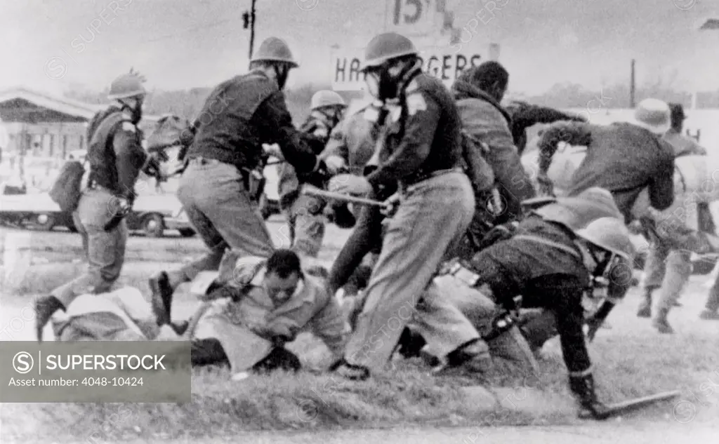 SNCC leader John Lewis (light coat in center) cringes as an Alabama State Trooper swings his club at Lewis' head. Lewis was hospitalized for a head injury. March 7, 1965.