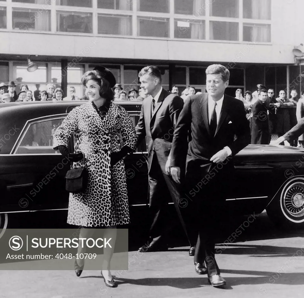 Escorted by President Kennedy, Jacqueline Kennedy departs for trip to India and Pakistan wearing an Oleg Cassini leopard skin coat. She touched off a fashion demand that is estimated to have cost over 250,000 leopards death, nearly wiping out the species. Mar. 8, 1962.