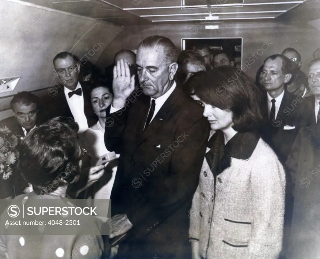 Johnson taking oath. Ladybird Johnson and former first lady Jacqueline Kennedy witness U.S. District Judge Sarah T. Hughes administering the oath of office to Lyndon B. Johnson in the conference room aboard Air Force One at Love Field, Dallas, Texas.  Photographer Cecil Stoughton. November 22, 1963.