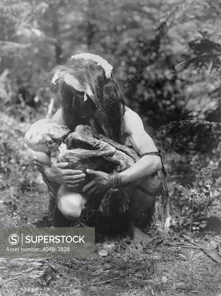 Kwakiutl man, crouched, cradling mummy bound in fetal position, title: Preparing to eat the mummy', photograph by Edward S. Curtis, circa 1911.