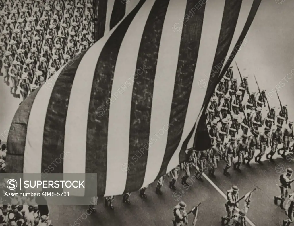 Dramatic photo of US flag and uniformed soldiers carrying rifles with bayonets fixed, marching in a parade during national mobilization in 1942.