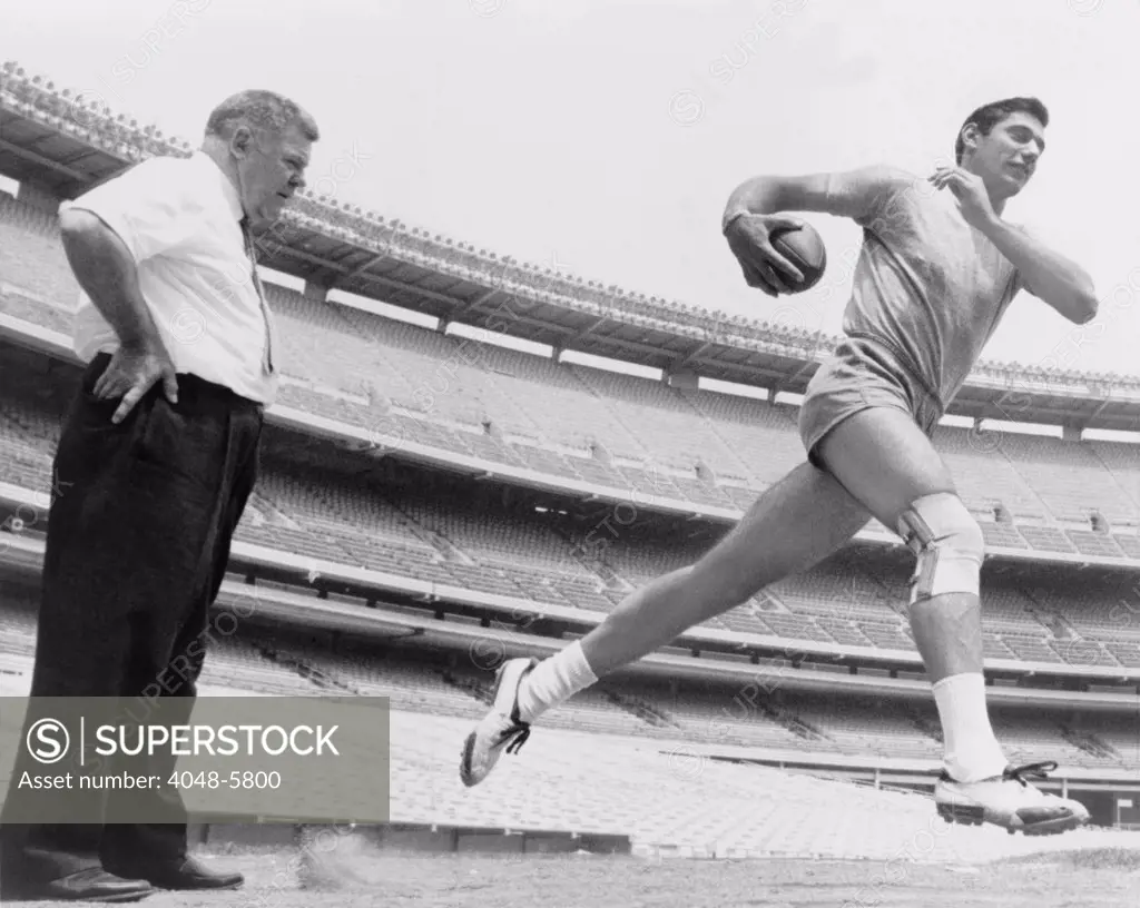 Joe Namath (b. 1943), quarterback with the New York Jets, running during light workout to test his injured knee, as coach Weeb Eubank observes. After his football career, Namath had knee replacement surgery. 1965.