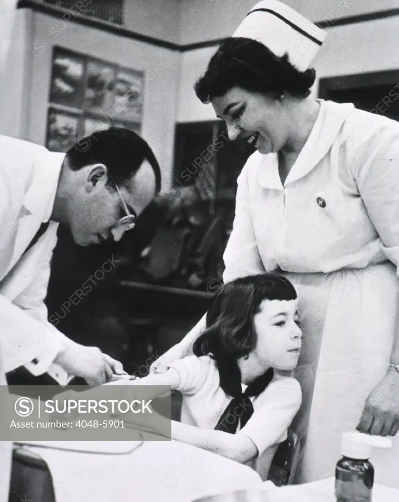 Jonas E. Salk (1914-1995), American physician who developed the first effective Polio vaccine inoculating child as nurse stands by. Ca. 1955.