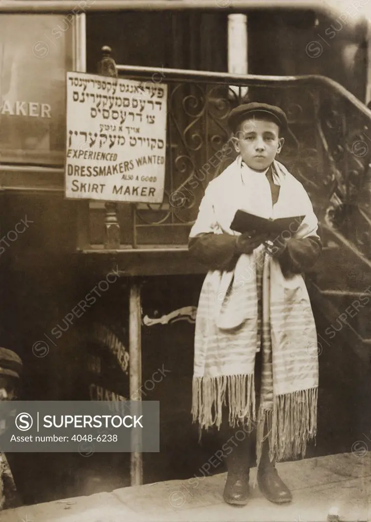 Jewish boy wearing a prayer shawl and boy reading from a Hebrew Bible on Yom Kippur, on a New York City street in a Jewish neighborhood. Ca.1911.