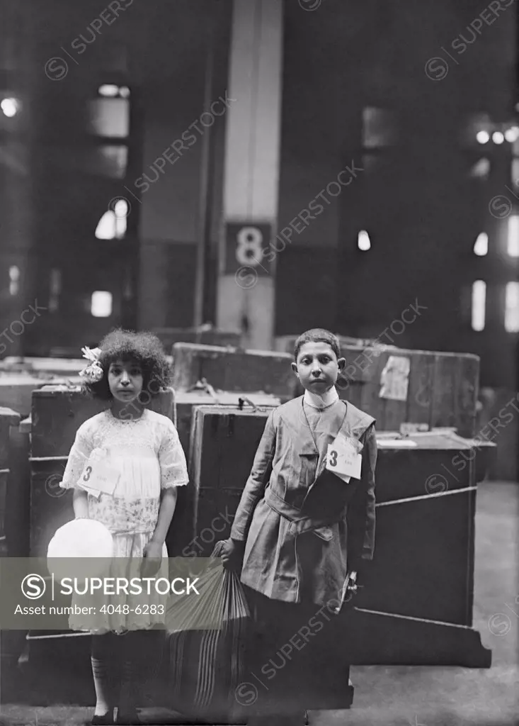 Two well dressed immigrant children tagged with 'ERIE' labeled at Ellis Inland, in front of many steamer trunks. Ca. 1910.