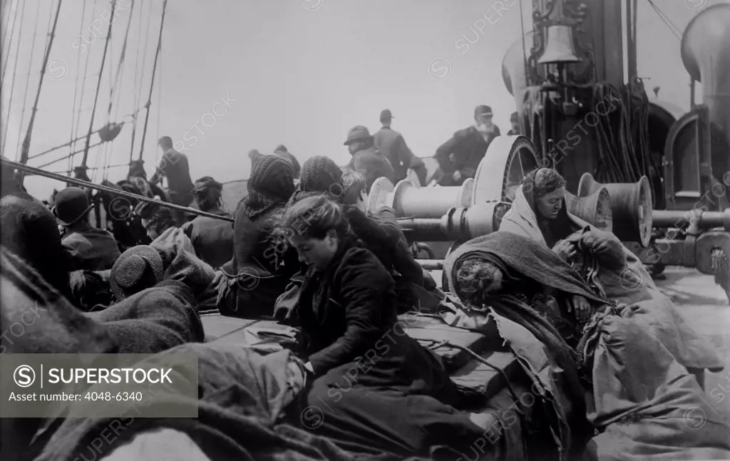A young women reading on the steerage deck of an ocean liner approaching the Port of New York. Other women passengers, wrapped in blankets, sleep amid the hardware of the ship's equipment. Ca. 1910.