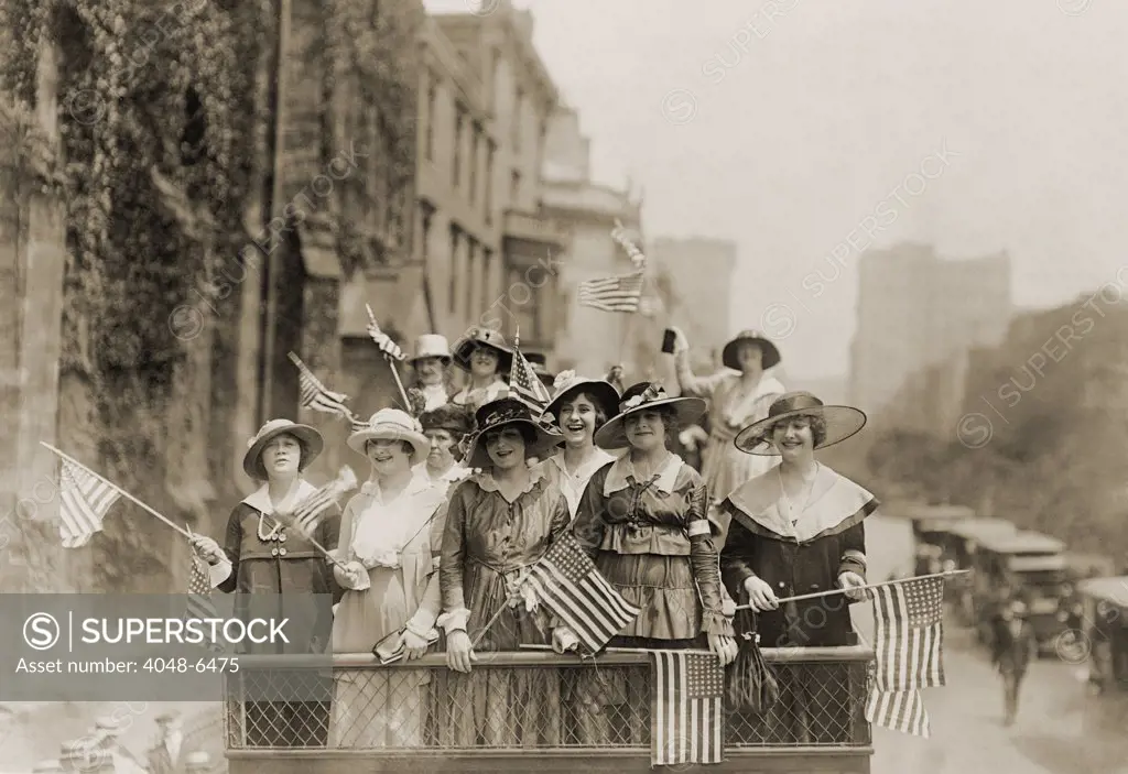Smiling young women waving flags from the top of an open vehicle in a New York City parade. Ca. 1910-1915.