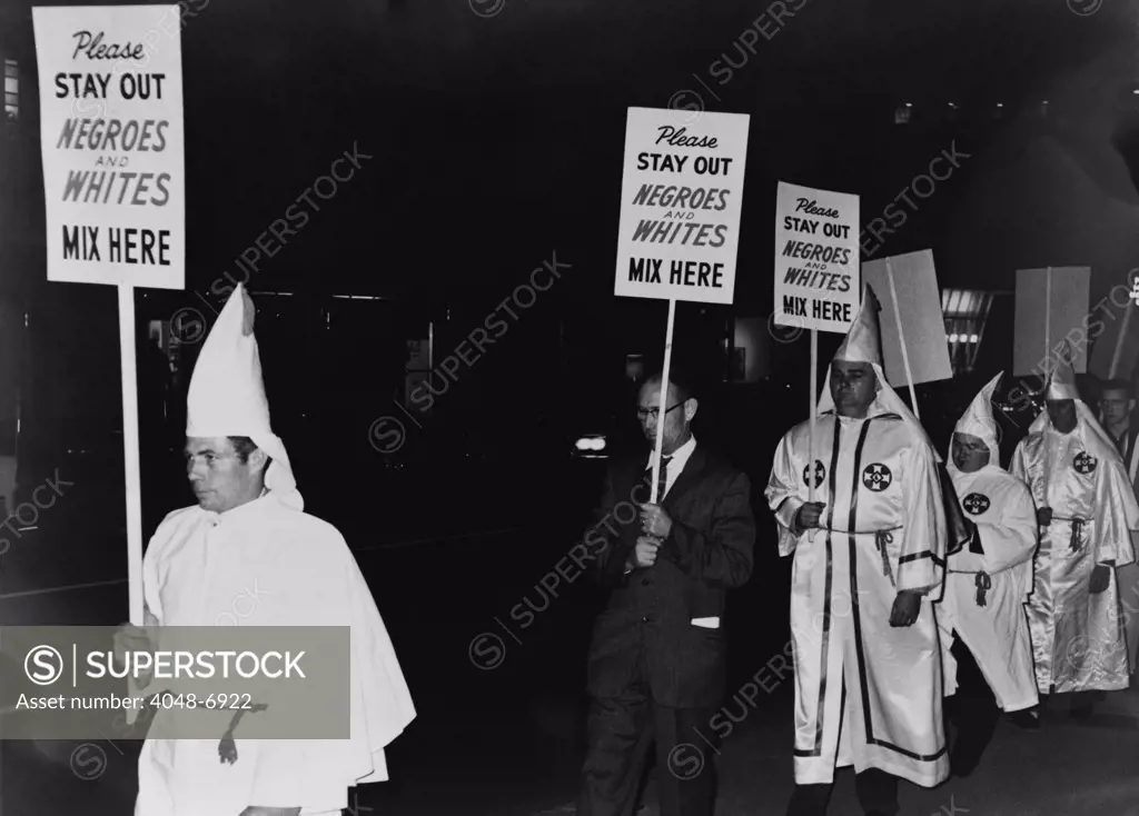 Ku Klux Klan members, in hooded white robes hoods, picket an integrated business carrying placards stating, 'Please stay out, Negroes and Whites mix here'. 1964.