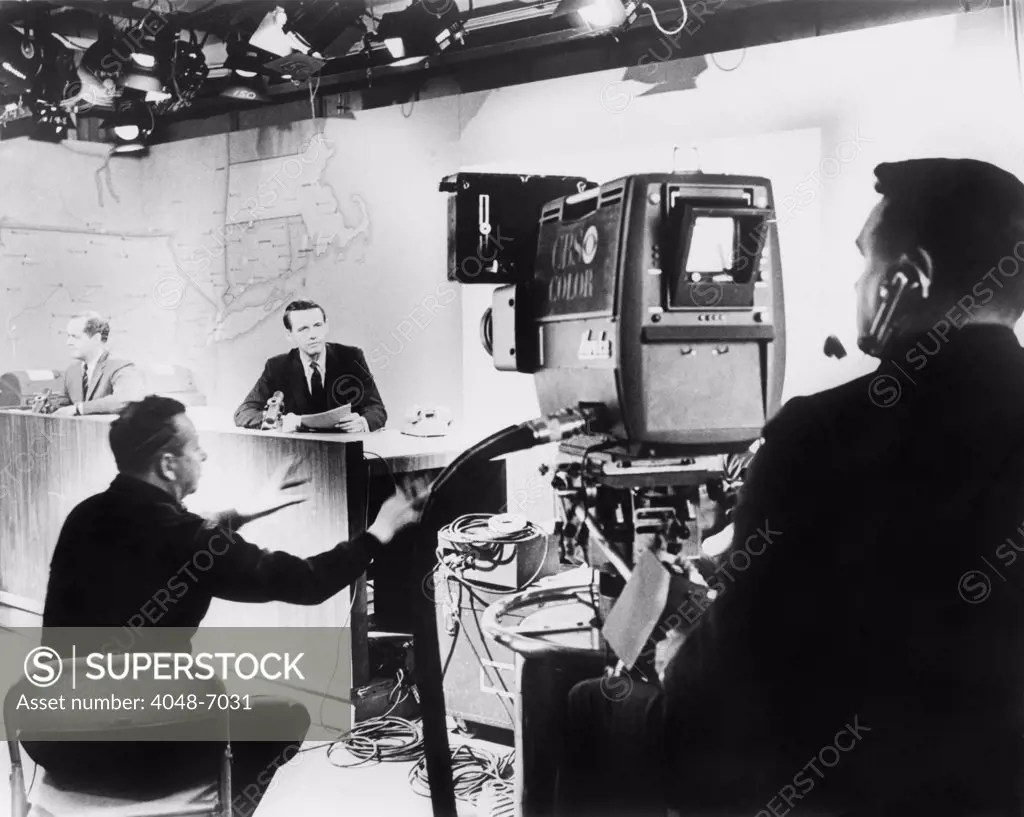 Under hot studio lights, a CBS News anchor performs under the director of his producer (seated between anchor and camera), for a color television camera in 1967.