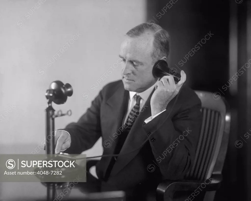 Businessman dialing an early 20th century desk phone with separate ear receiver and mouth transmitter. Ca. 1920.