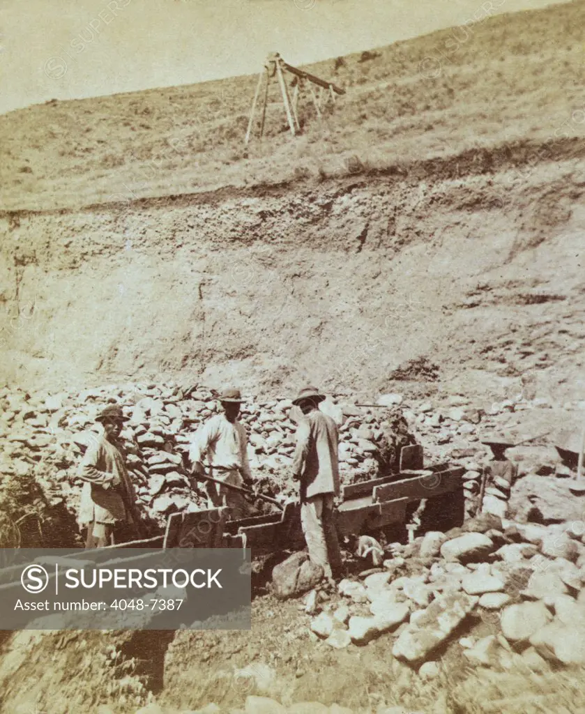 Chinese prospectors washing gold in a sluice box placed in the stream to channel water flown. Gold-bearing soil is placed at the top of the box, which is lined with horizontal riffles that cause the gold to drop out of flowing liquid. 1871 photo by William Henry Jackson, probably taken in Colorado, Nevada or Arizona, during the Wheeler expedition.
