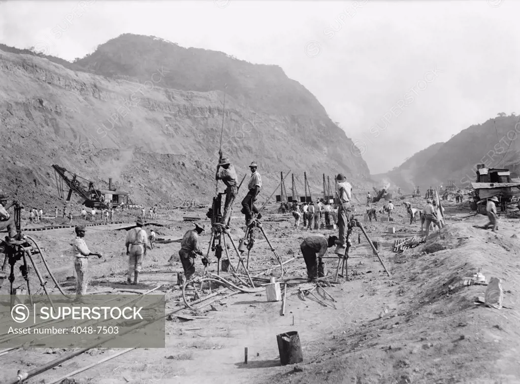 Panama Canal construction showing workers drilling holes for dynamite in bedrock, as they cut through the mountains of the Isthmus. Steam shovels in the background move the rubble to railroads car. 1913.