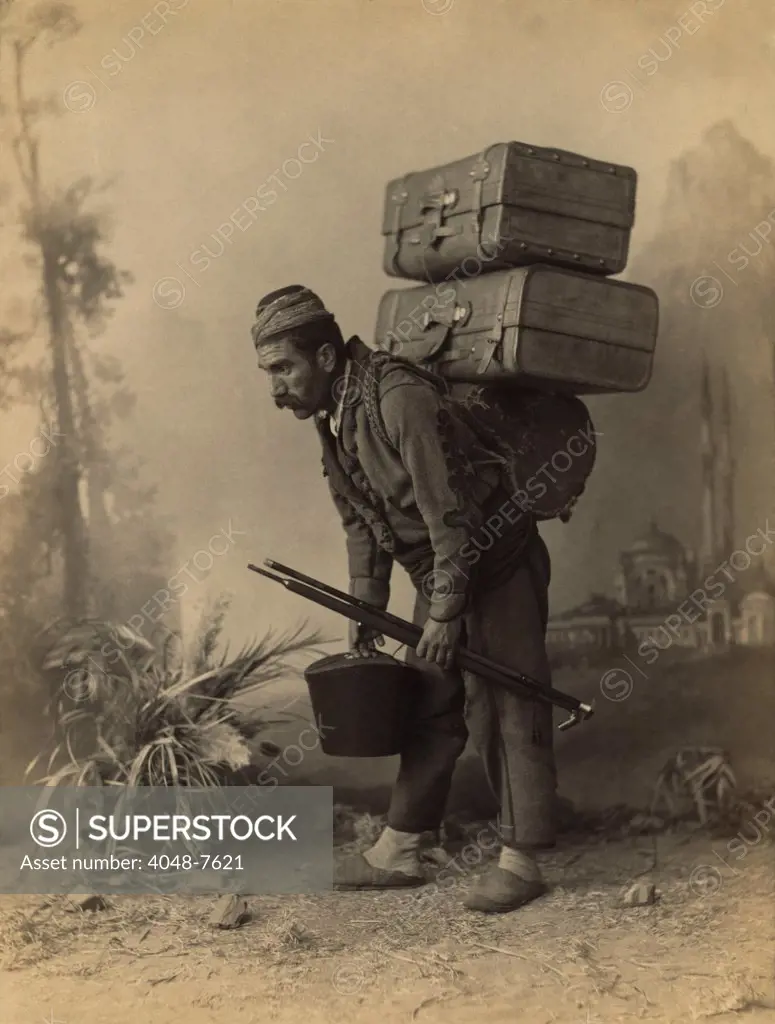 Turkish porter carrying luggage on his back. Studio portrait with painted backdrop of landscape with mosque, by Abdullah Freres, ca. 1890.